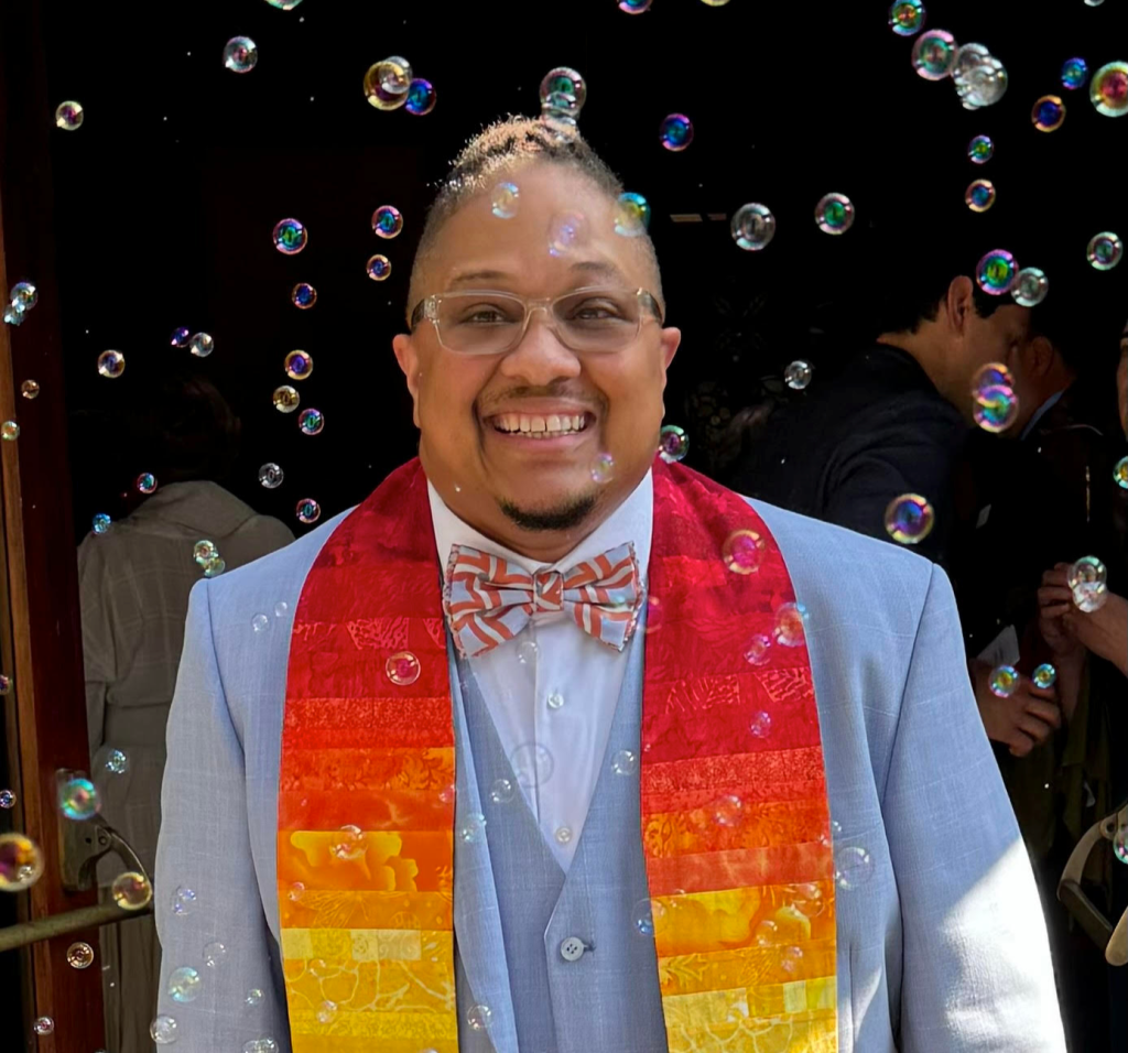 Mykal, a Black man, smiles broadly, wearing glasses, a rainbow stole, a light blue jacket and vest, and a snazzy bow tie, surrounded by falling bubbles